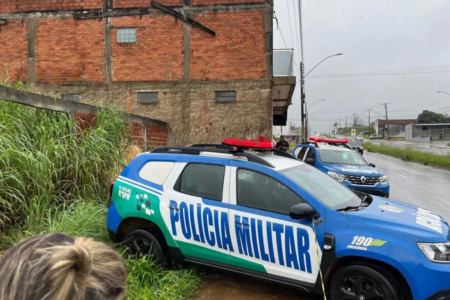 Imagem colorida mostra uma viatura da Polícia Militar parada em uma área com mato alto, em frente a uma casa com parede de tijolos.