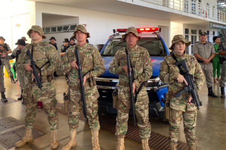 Imagem colorida mostra quatro policias do Batalhão Rural em frente a uma viatura da Polícia. Todos estão com armas nas mãos.