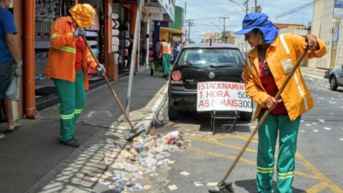 Imagem colorida mostra dois trabalhadores da Comurg varrendo uma rua em Goiânia cheia de lixo e papéis.