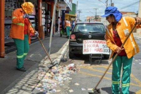 Imagem colorida mostra dois trabalhadores da Comurg varrendo uma rua em Goiânia cheia de lixo e papéis.