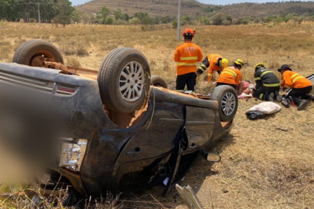 Imagem colorida mostra um carro preto de cabeça para baixo após capotar em uma área de mata.