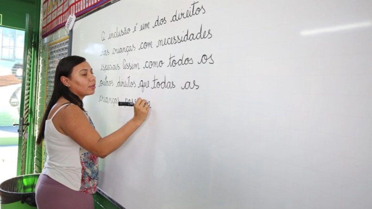 Imagem mostra uma mulher, professora, escrevendo com pincel em um quadro branco