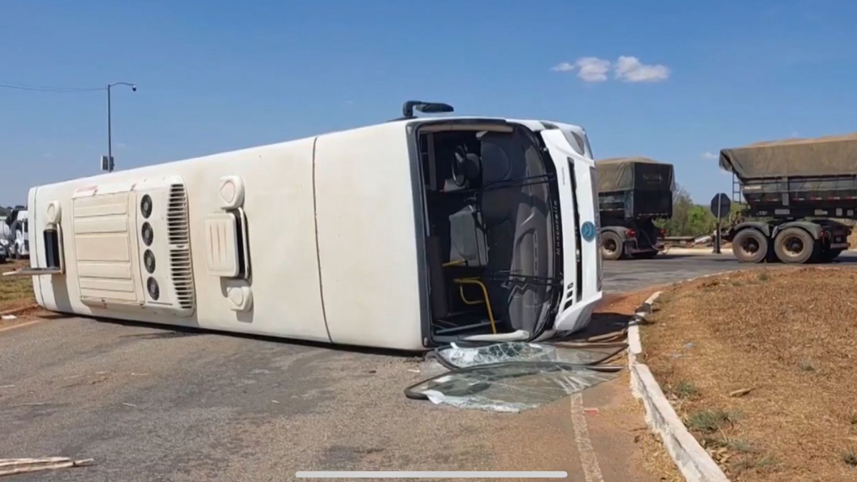 Imagem colorida mostra um ônibus branco tombado em uma rodovia.