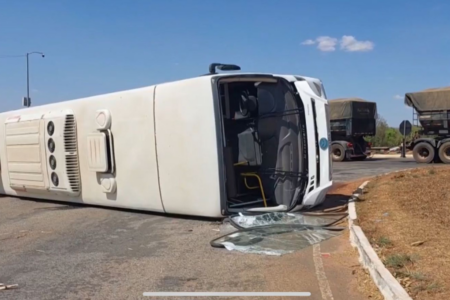 Imagem colorida mostra um ônibus branco tombado em uma rodovia.