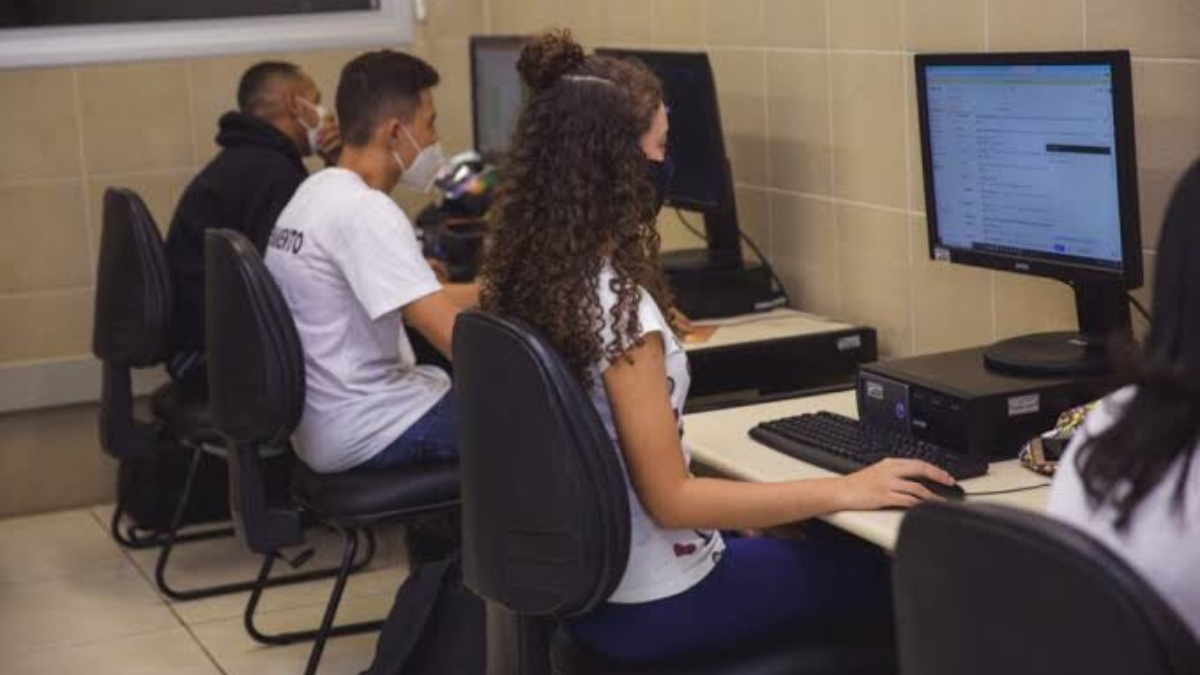 Imagem colorida mostra dois jovens, um menino e uma menina, virados de costas e mexendo no computador.