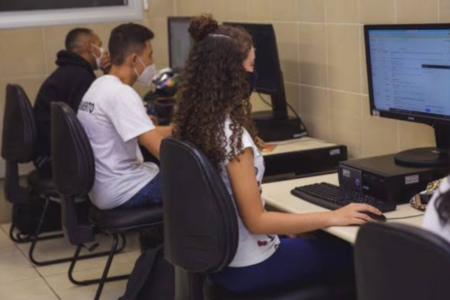 Imagem colorida mostra dois jovens, um menino e uma menina, virados de costas e mexendo no computador.