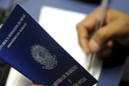 Imagem colorida mostra uma cartera de trabalho sendo segurada aberta e uma outra mão escrevendo em uma folha em branco.