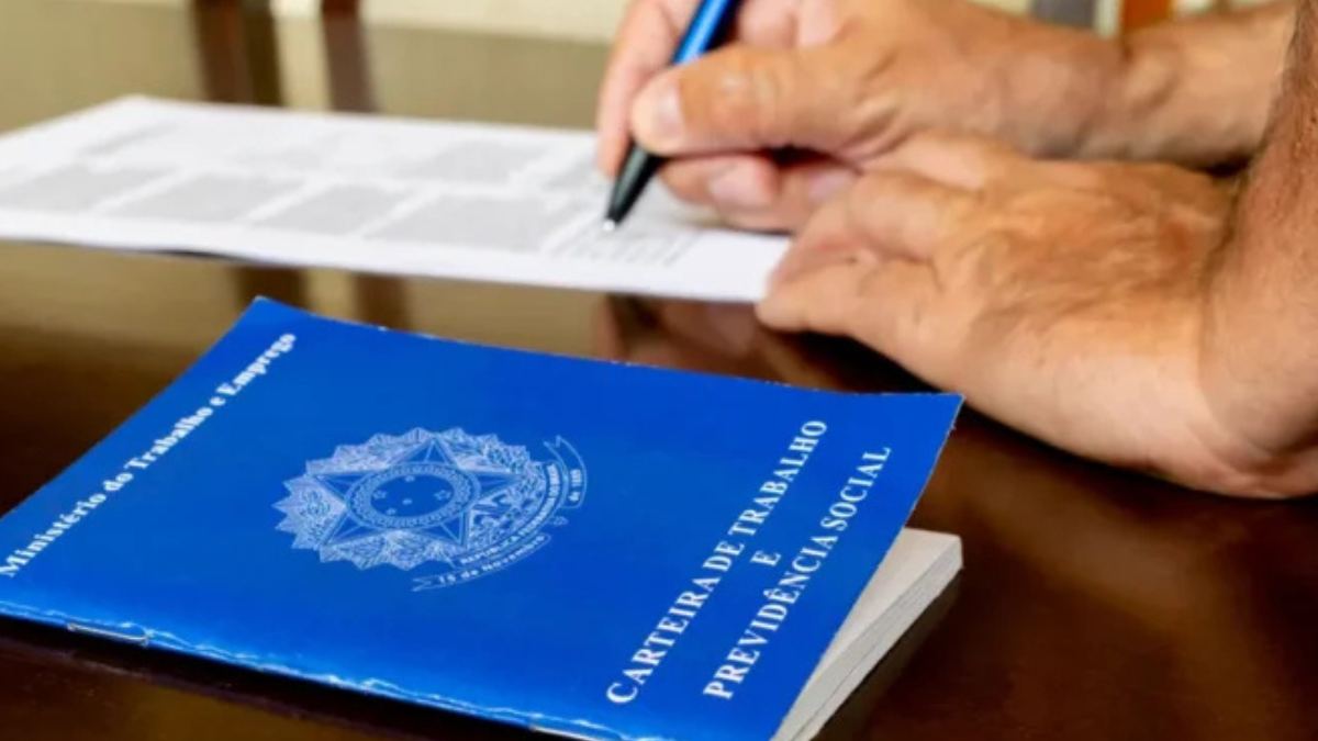 Imagem colorida mostra uma carteira de trabalho em primeiro plano e, em segundo, a mão de uma pessoa escrevendo em um papel.