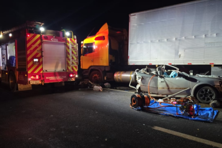 Imagem colorida mostra uma viatura dos Bombeiros ao lado de um caminhão carreta e um carro prata completamente amassado após um acidente.