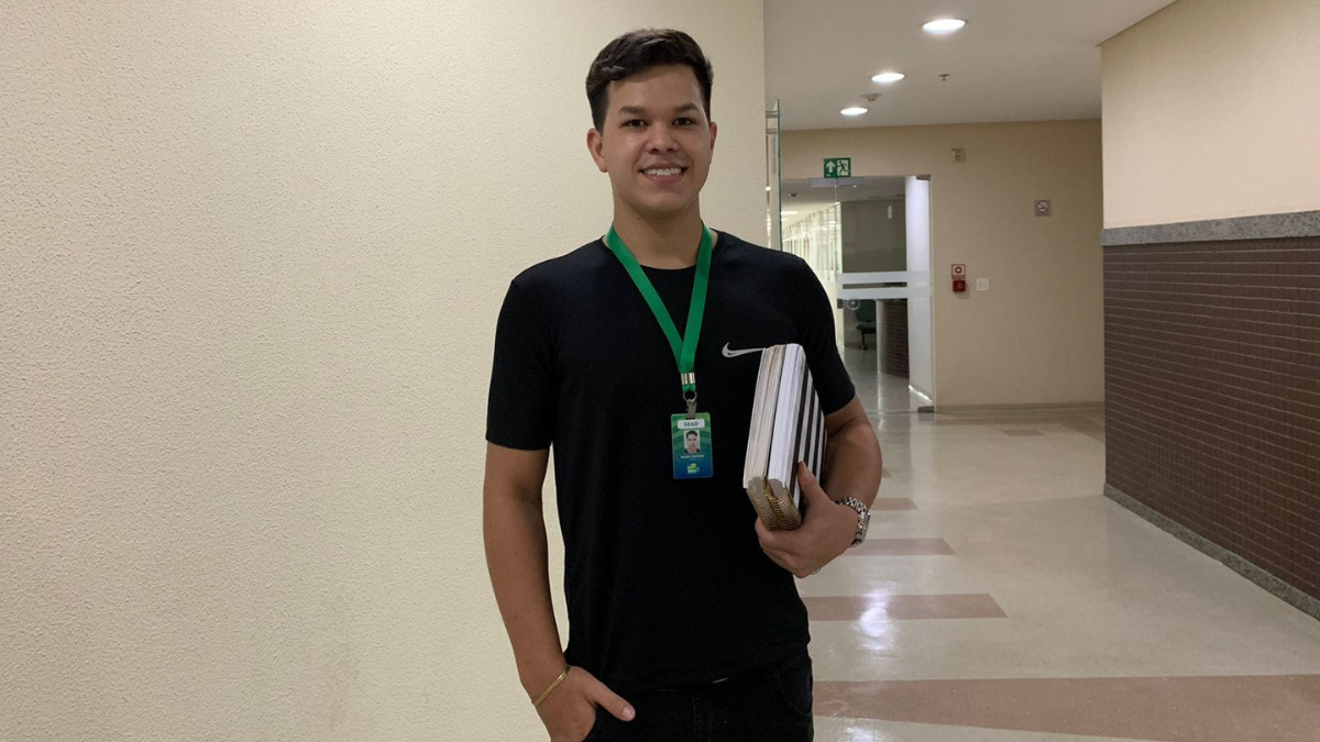 Imagem colorida mostra um homem em pé usando uma camiseta preta e um crachá. Ele também segura cadernos nos braços.