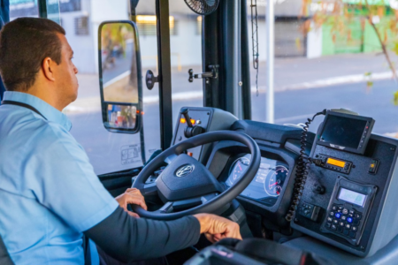 Imagem colorida mostra um motorista dentro de um ônibus do transporte coletivo.