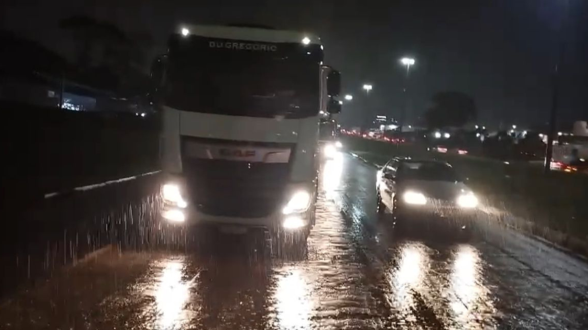 Imagem colorida mostra um caminhão e um carro de passeio parados na rodovia molhada pela chuva.