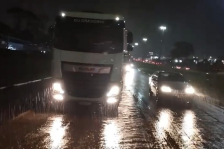 Imagem colorida mostra um caminhão e um carro de passeio parados na rodovia molhada pela chuva.