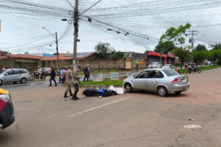 Imagem colorida mostra um veículo prata virando no cruzamento de duas ruas e uma moto caída no mesmo local.