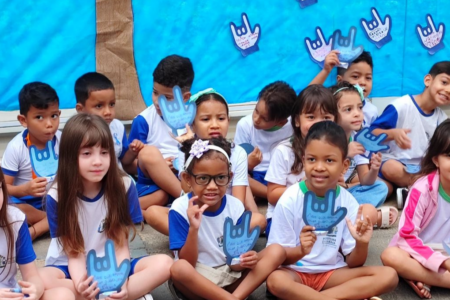 Imagem colorida mostra diversas crianças usando uniforme escolar sentadas em frente de um painel com desenhos de língua de sinais.