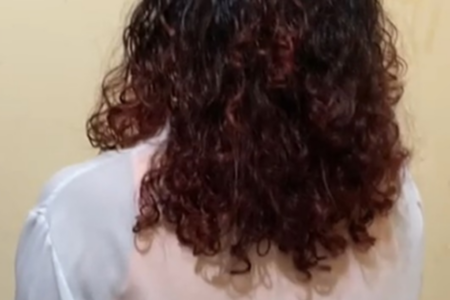 Imagem colorida mostra uma menina de costas. Na foto aparece só o cabelo dela, cacheado, e uma parte de sua camiseta branca.