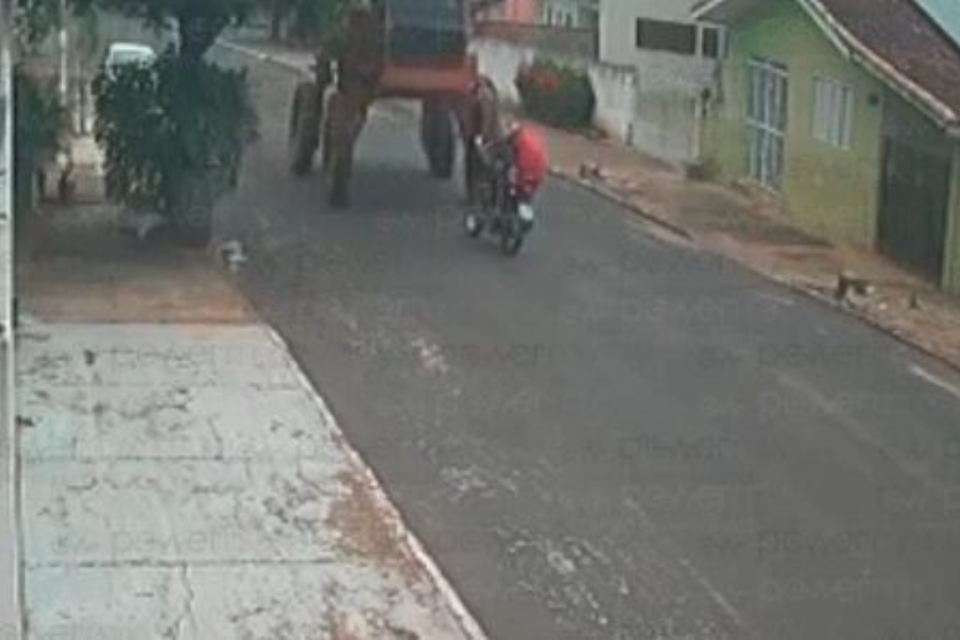 Imagem colorida mostra uma moto, com duas pessoas, em uma rua, e uma máquina agrícola vindo no sentido oposto antes de um acidente acontecer.