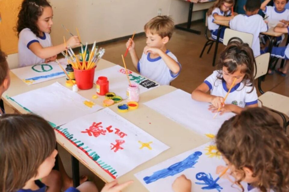 A imagem mostra crianças em uma creche realizando tarefas pedagógicas de pintura