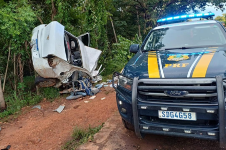 Imagem colorida mostra uma viatura da PRF em primeirp plano e do lado esquerdo, um carro branco totalmente destruído após se chocar com um caminhão em uma rodovia.