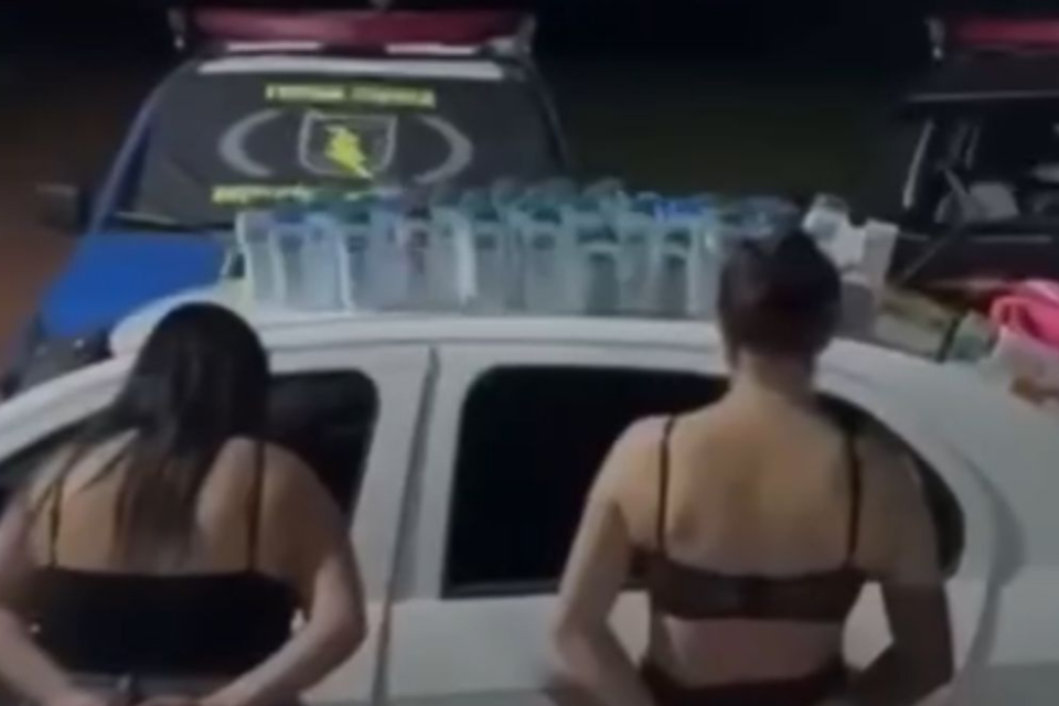 Imagem colorida mostra duas mulheres de costas ao lado de uma viatura da polícia.