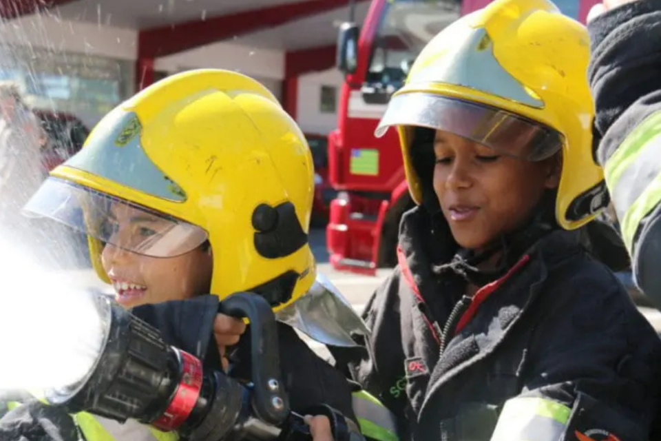 Imagem colorida mostra dois meninos vestidos como bombeiros, usando a roupa preta e capacete amarelo.