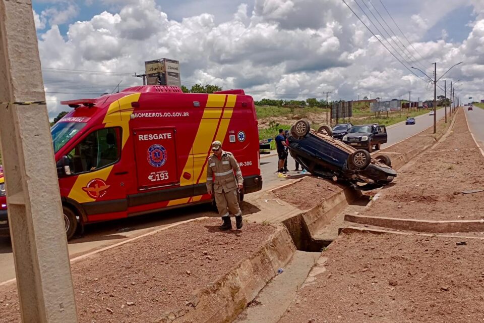 A foto mostra um carro capotado, virado com os pneus para cima, no meio de um canteiro central. A frente uma ambulância do Corpo de Bombeiros realizando o resgate da vítima