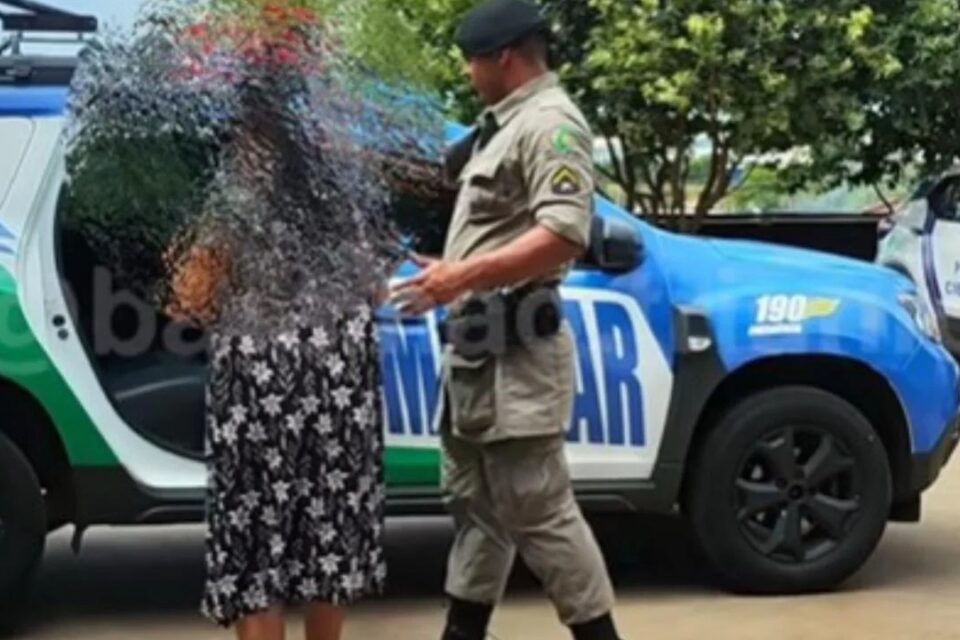 A imagem mostra uma foto onde uma mulher é conduzida para uma viatura da Polícia Militar por um agente