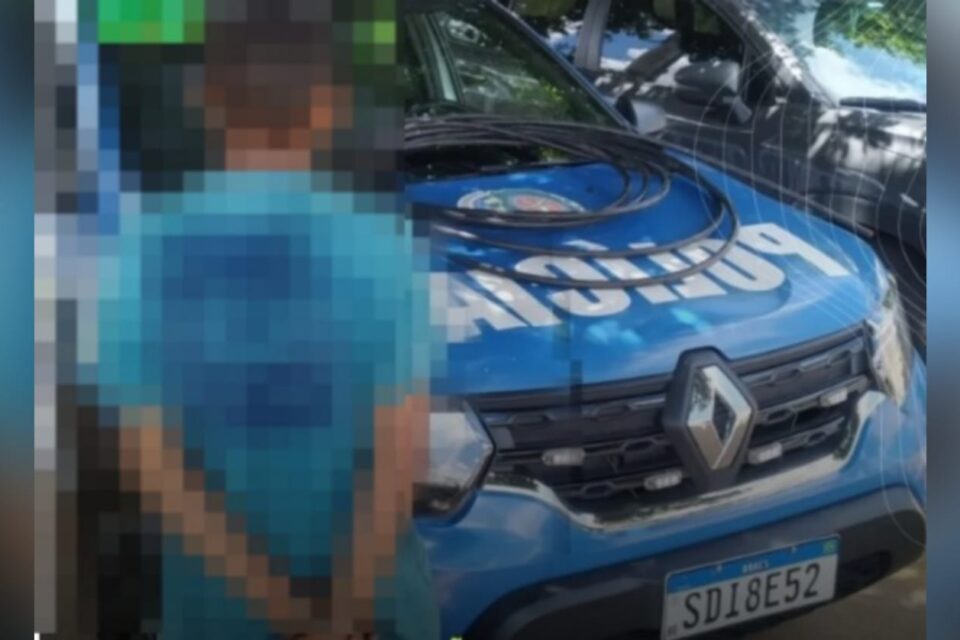 A imagem mostra uma foto com o supeito de furtar cabos de costas e em frente uma viatura da PM