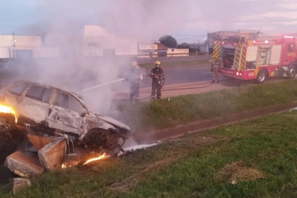 Imagem colorida mostra um carro pegando fogo e bombeiros apagando as chamas à beira de uma rodovia.