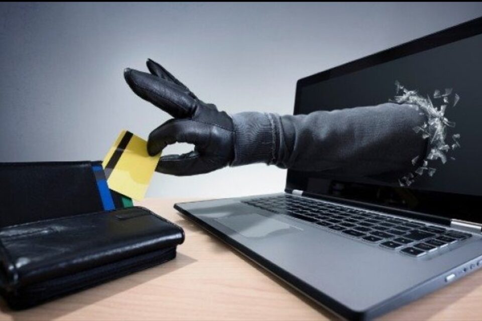 A imagem mostra um braço saindo da tela de um notebook e retirando um cartão de uma carteira sobre uma mesa de madeira
