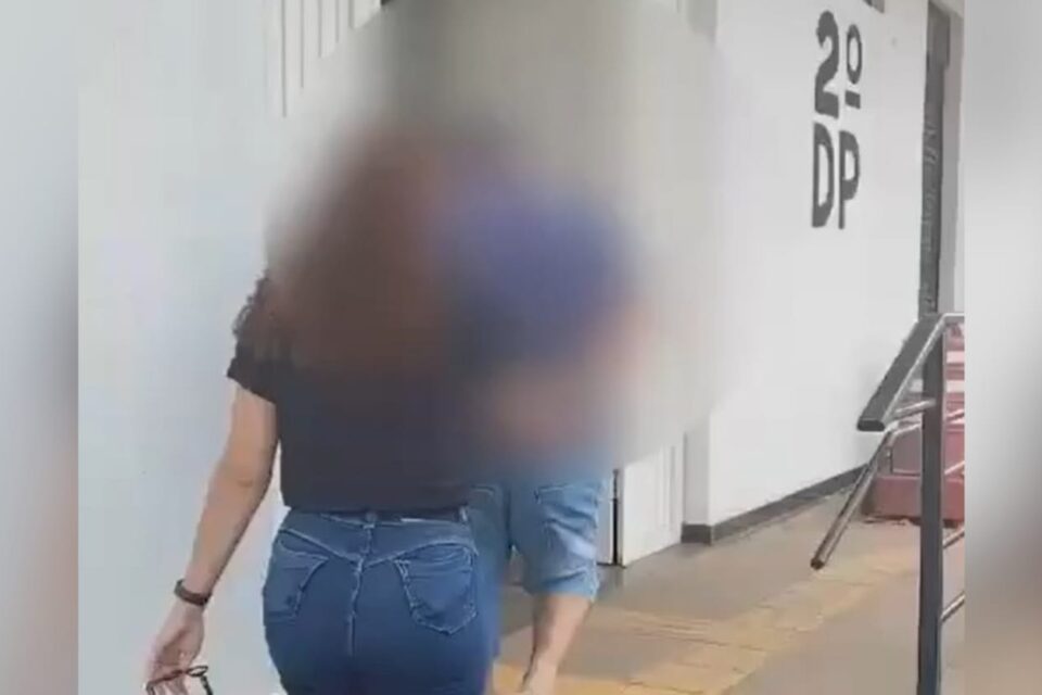 A imagem mostra um homem, suspeito de estelionato, algemado e sendo conduzido por uma policial até a delegacia