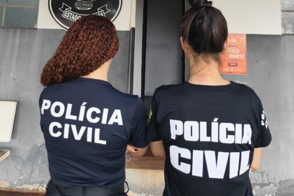 A imagem mostra duas agentes da Polícia Civil de costas de frente para entrada da delegacia
