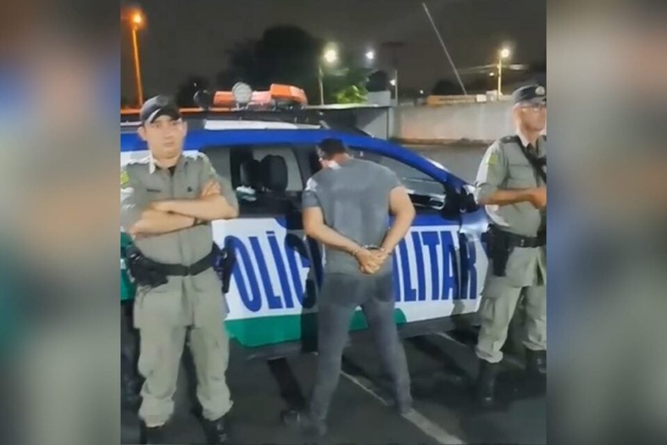 A imagem mosta um homem suspeito de tráfico de drogas preso em frente a uma viatura da Polícia Militar e ao lado dele, dois policias