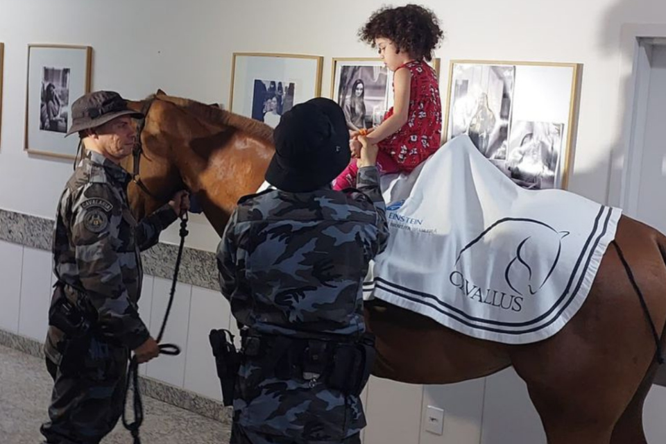 Imagem colorida mostra uma criança em cima de um cavalo e um policial ao lado auxiliando.