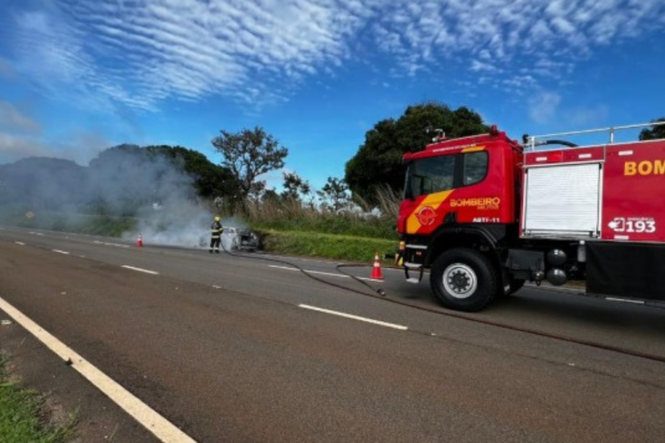 Imagem colorida mostra um caminhão dos bombeiros em uma rodovia atrás de um carro em chamas após pegar fogo.