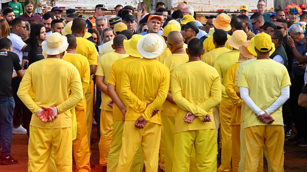 Imagem colorida mostra um grupo de detentos virados de costas. Todos estão usando uma roupa totalmente amarela.