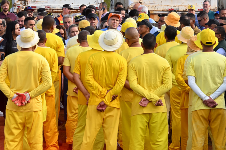 Imagem colorida mostra um grupo de detentos virados de costas. Todos estão usando uma roupa totalmente amarela.