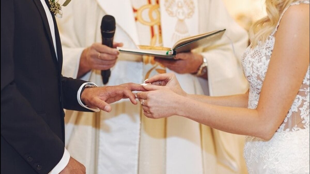 Foto ilustrativa de um casal em frente a um padre em uma cerimonia de casamento, na foto é mostrado o momento da troca de alianças do casal