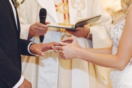 Foto ilustrativa de um casal em frente a um padre em uma cerimonia de casamento, na foto é mostrado o momento da troca de alianças do casal