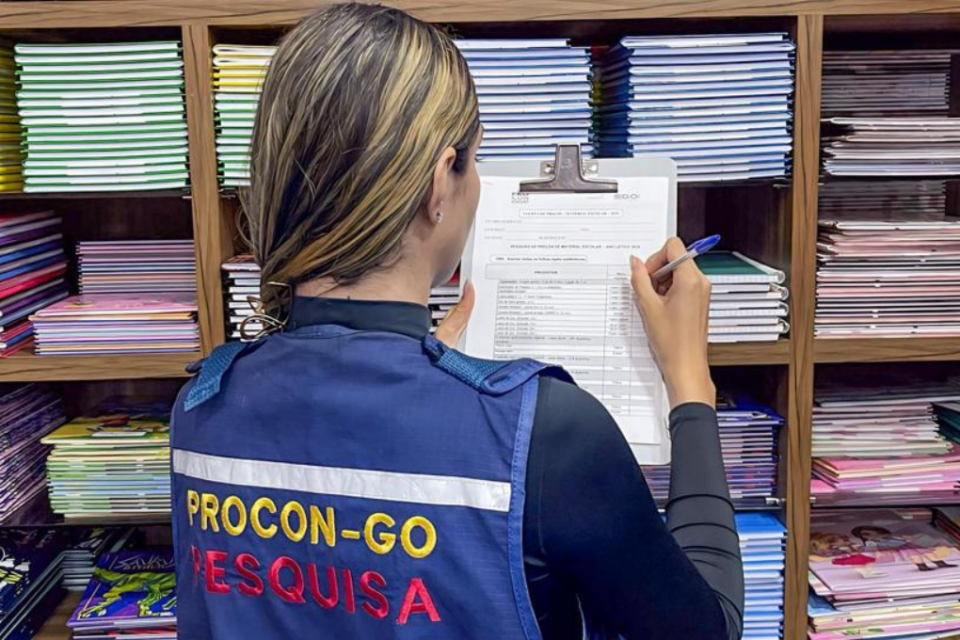 Imagem colorida mostra uma fiscal do Procon Goiás com uma prancheta na mão olhando para uma prateleira com materiais escolares.