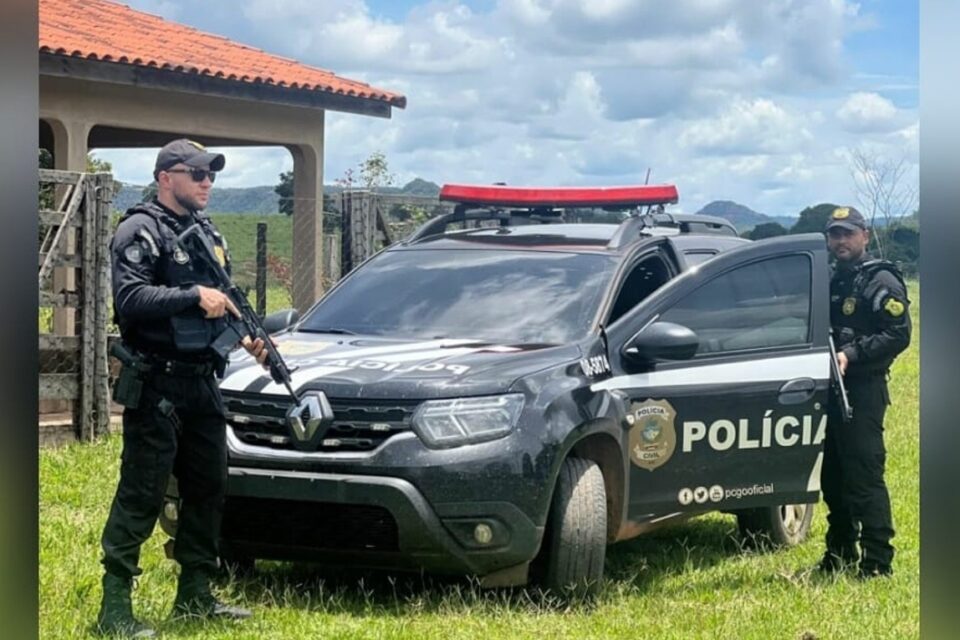 A imagem mostra uma viatura parada em um propriedade onde foi cumprido mandado de busca e apreensão. Um suspeito foi preso. Ao lado da viatura estão dois policiais civis, um deles segura uma arma