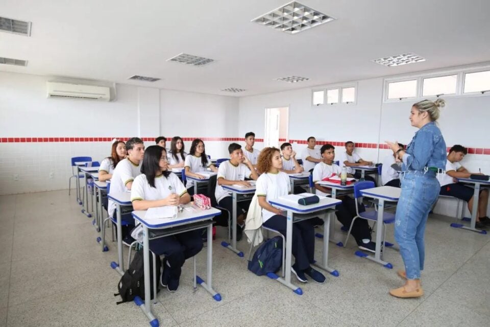 A imagem mostra uma sala de aula de um colégio estadual onde uma professora está ministrando aula para os alunos