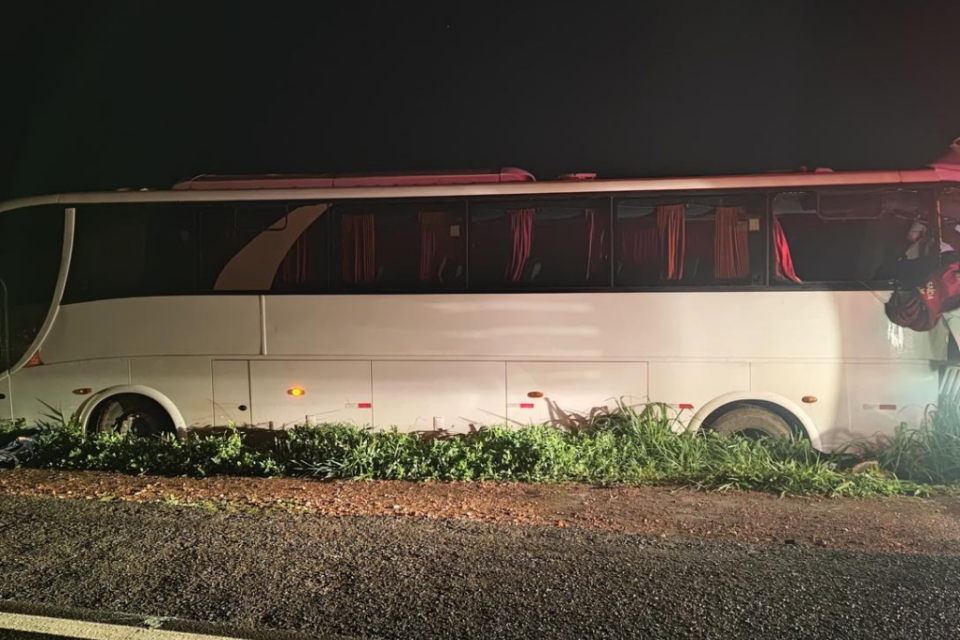Imagem colorida mostra um ônibus branco parado no canteiro de uma rodovia após um acidente.