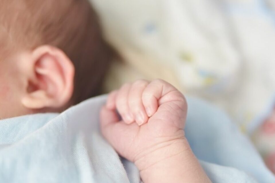 A imagem mostra a mãozinha de um bebê deitado sobre uma cama