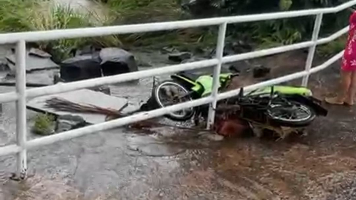 Imagem colorida mostra uma moto quase caindo em um córrego após ser arrastada pela chuva.