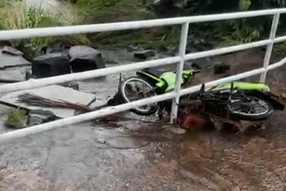 Imagem colorida mostra uma moto quase caindo em um córrego após ser arrastada pela chuva.