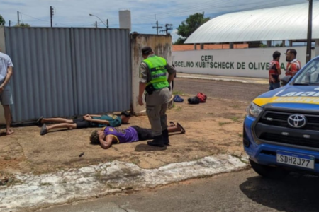 Imagem colorida mostra um policial militar em pé na calada em frente a dois rapazes suspeitos por um crime que estão detados no chão.