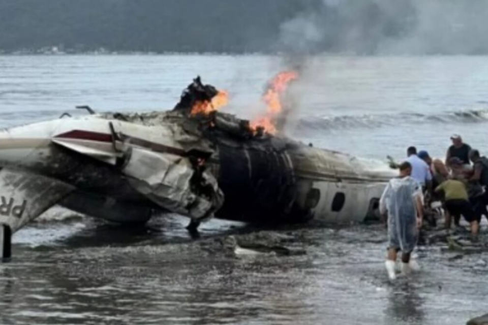 Imagem colorida mostra um avião após pegar fogo.. Ele está caído na beira do mar.