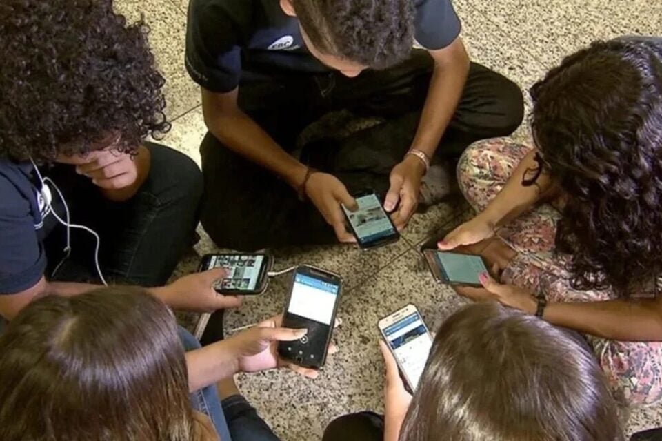 A imagem mostra alunos sentados em circulo usando celulares