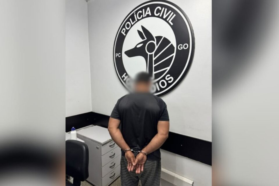 A imagem mostra um homem preso e algemado em uma delegacia